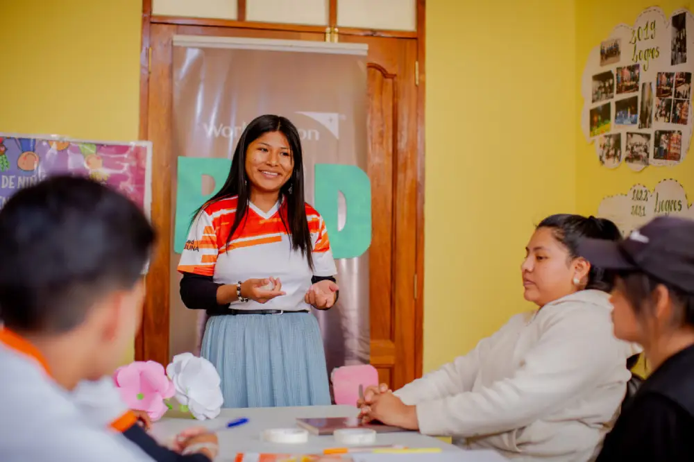 A girl stands at the end of a table, talking to a group of people sitting at the table.