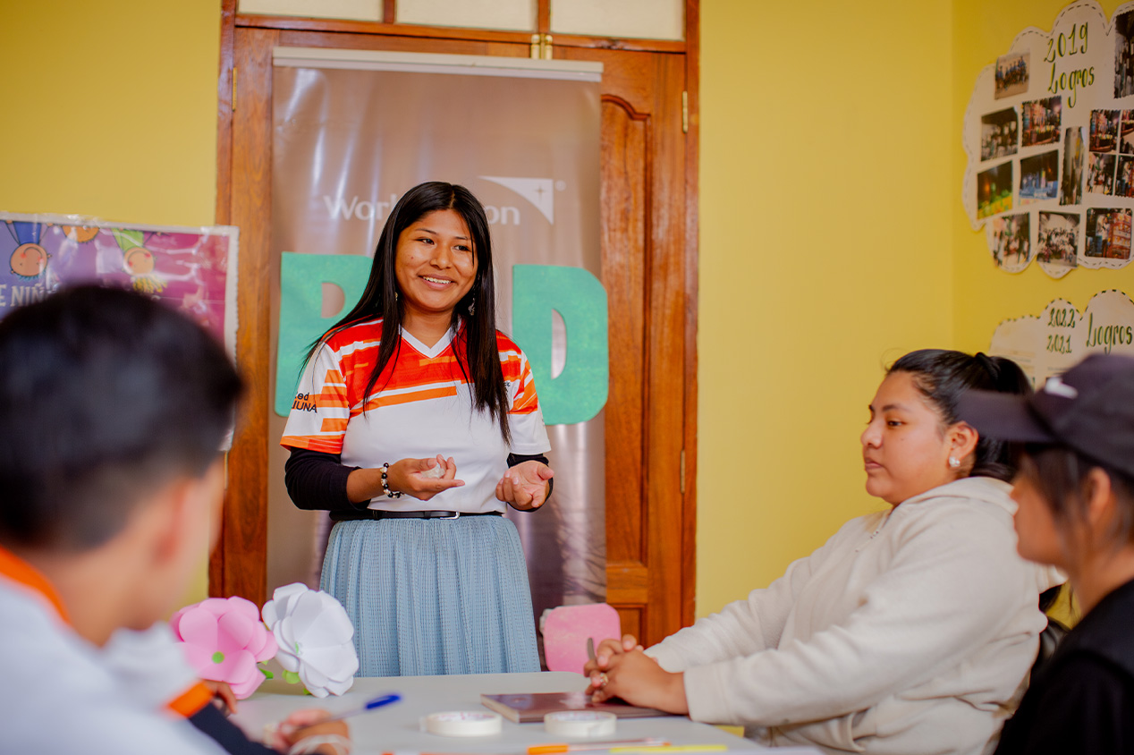 A girl stands at the end of a table, talking to a group of people sitting at the table.