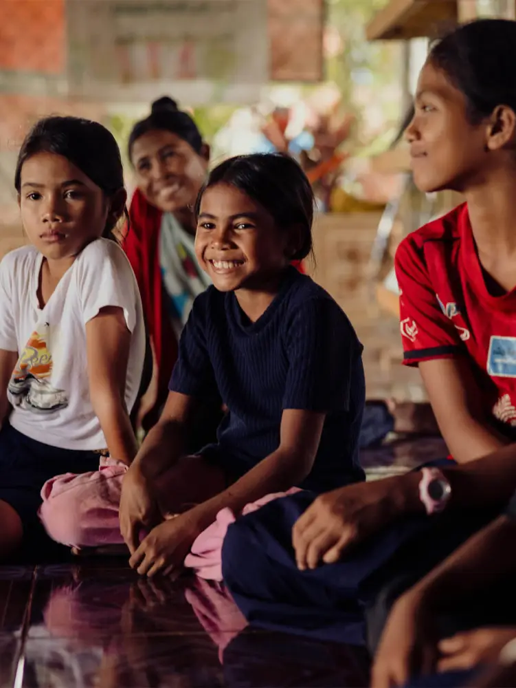 A young girl sits on the floor with a group of children, smiling.