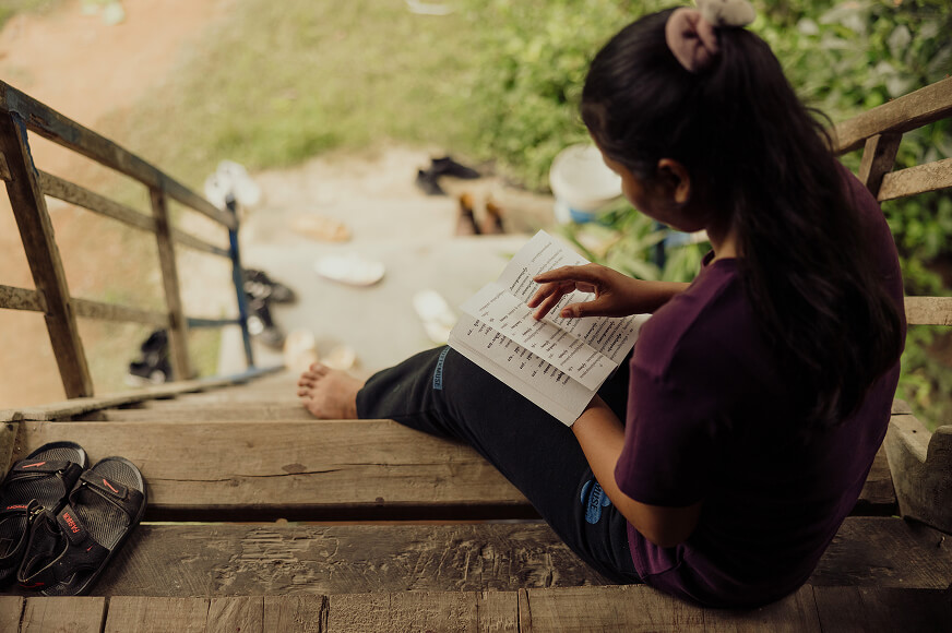 An adolescent girl sits on the front step of her hut in Cambodia, reading a book for her school studies. 