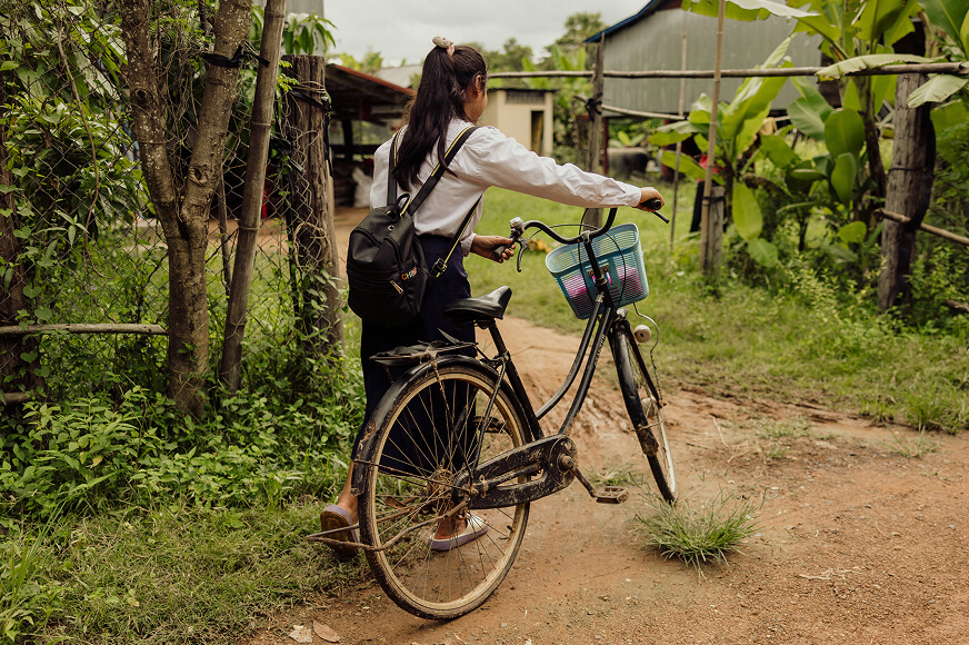 An adolescent girl is walking her bicycle on a rural Cambodian dirt road. She is wearing a school uniform and carrying a backpack. 