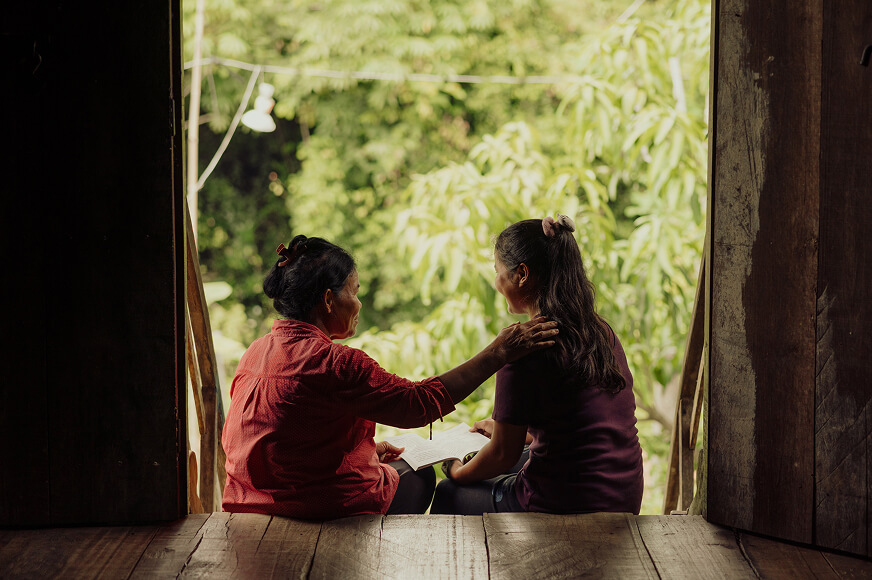 A mother and daughter sit at the entrance to their home. The mother has her arm on her daughter’s shoulder as they laugh and read a book together. 