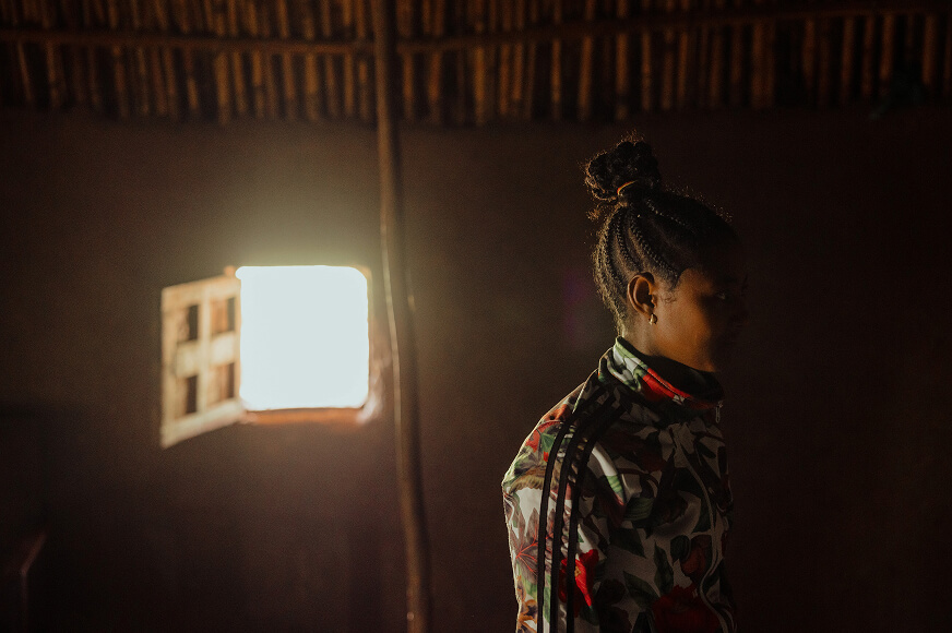 A woman sits in her Ethiopian hut with as light breaks through a little window. Her face is covered in shadow to protect her identity. 